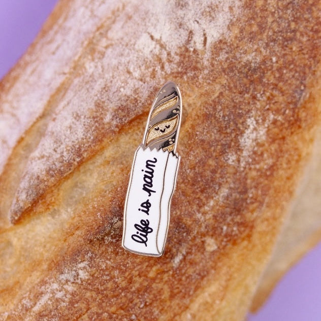 Bread with a 'Life is Pain' enamel pin on a purple background