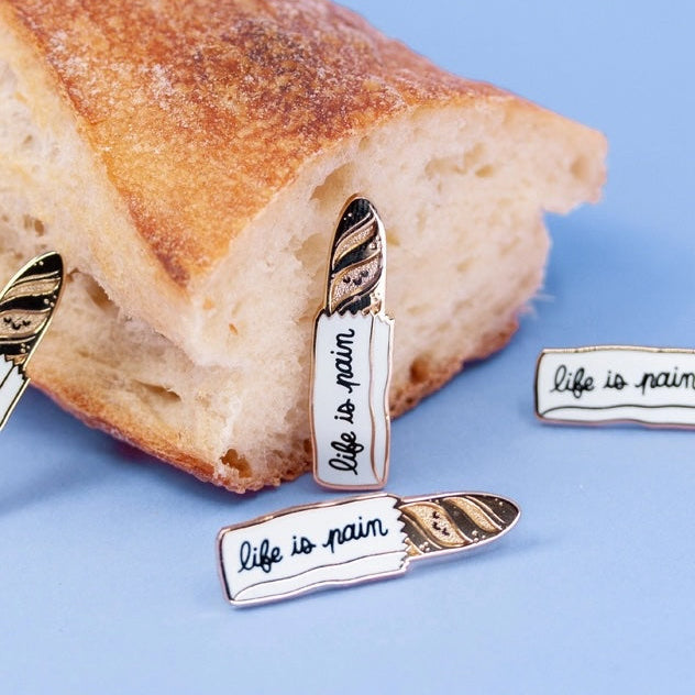 Enamel pins with 'life is pain' text on a piece of bread against a blue background