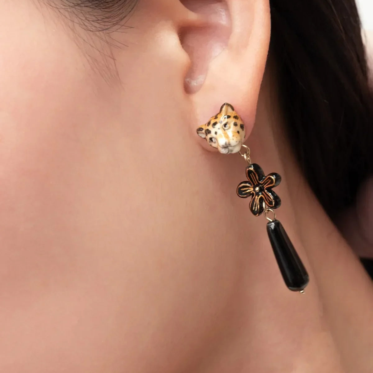 A close-up of a model's ear wearing a dangling earring with a gold leopard head stud, a black and gold flower charm, and a black teardrop drop.