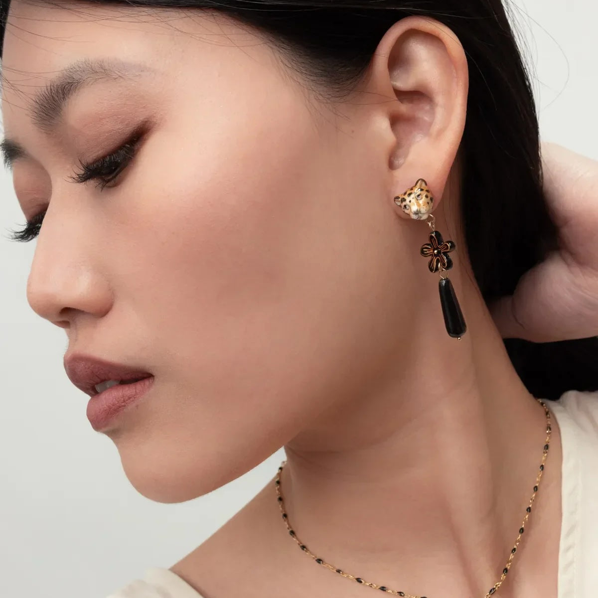 A close-up of a woman wearing a matching gold and black jewelry set, featuring a flower-shaped drop earring with a black teardrop pendant and a delicate beaded necklace with a flower charm.