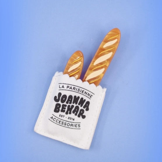 Two baguettes hair clips in a mini white microfiber bag with 'Joanna Behar' branding on a blue background