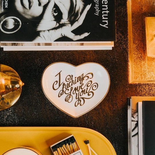 Heart-shaped coaster with 'I Fucking Love You' text on a table with books and candles.