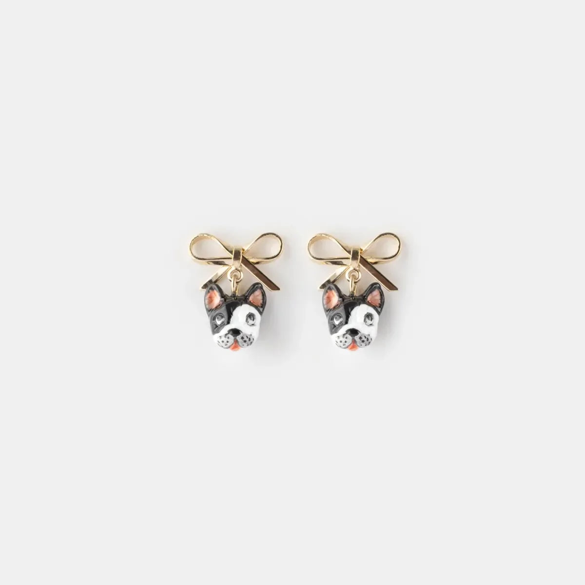 A pair of gold-tone stud earrings featuring small dog faces with black, white, and pink enamel details, tied with tiny gold bows above them, shown on a light gray background.