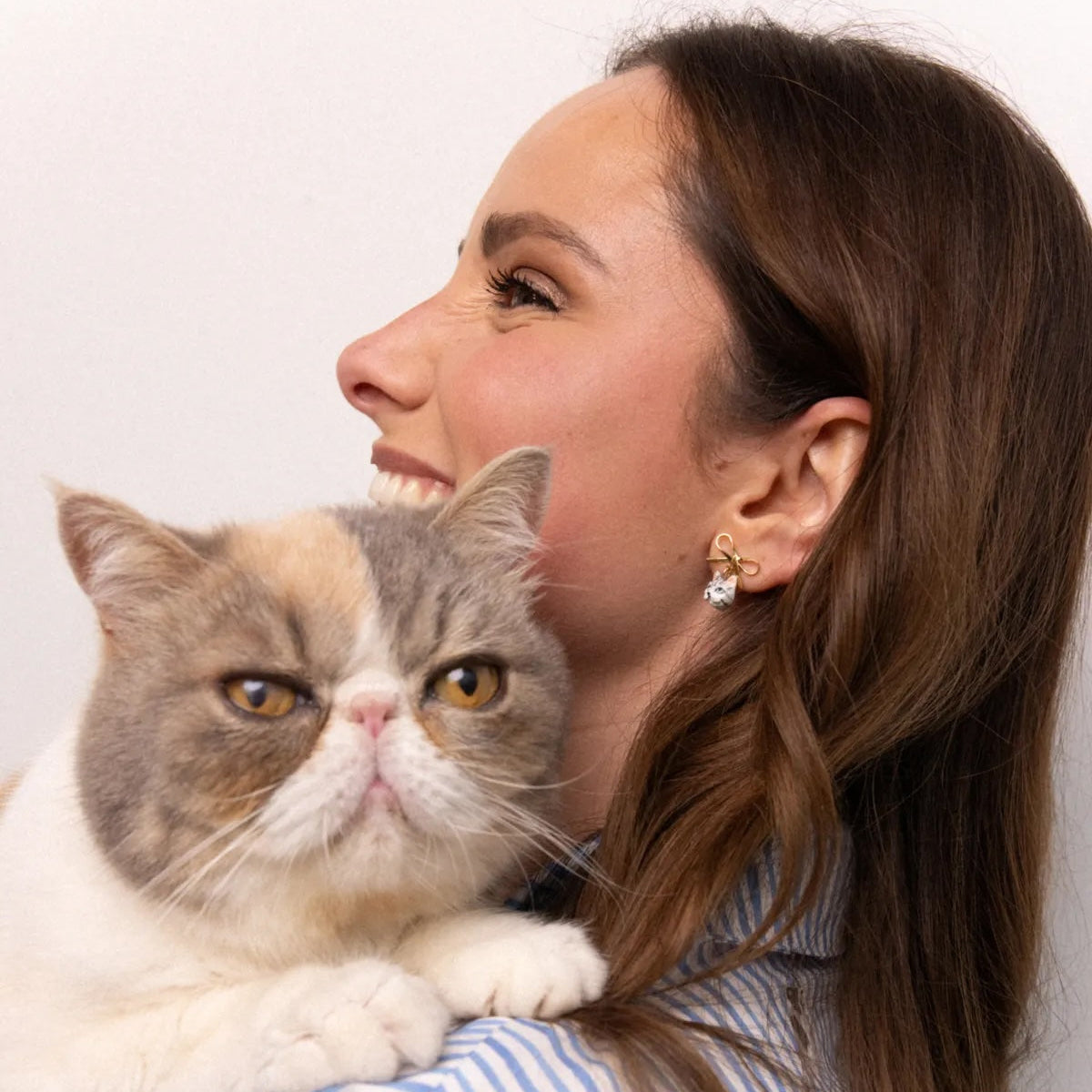 A woman with long brown hair smiles while holding a fluffy gray and white cat against a plain light background. She is wearing a blue and white striped shirt.