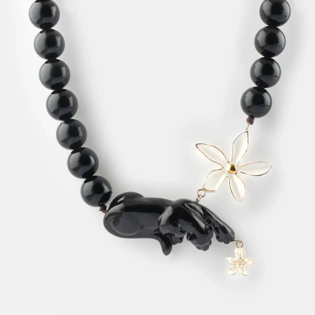 A black beaded necklace featuring a glossy black carved animal pendant, a white flower charm, and a small gold flower charm, displayed on a light gray background.