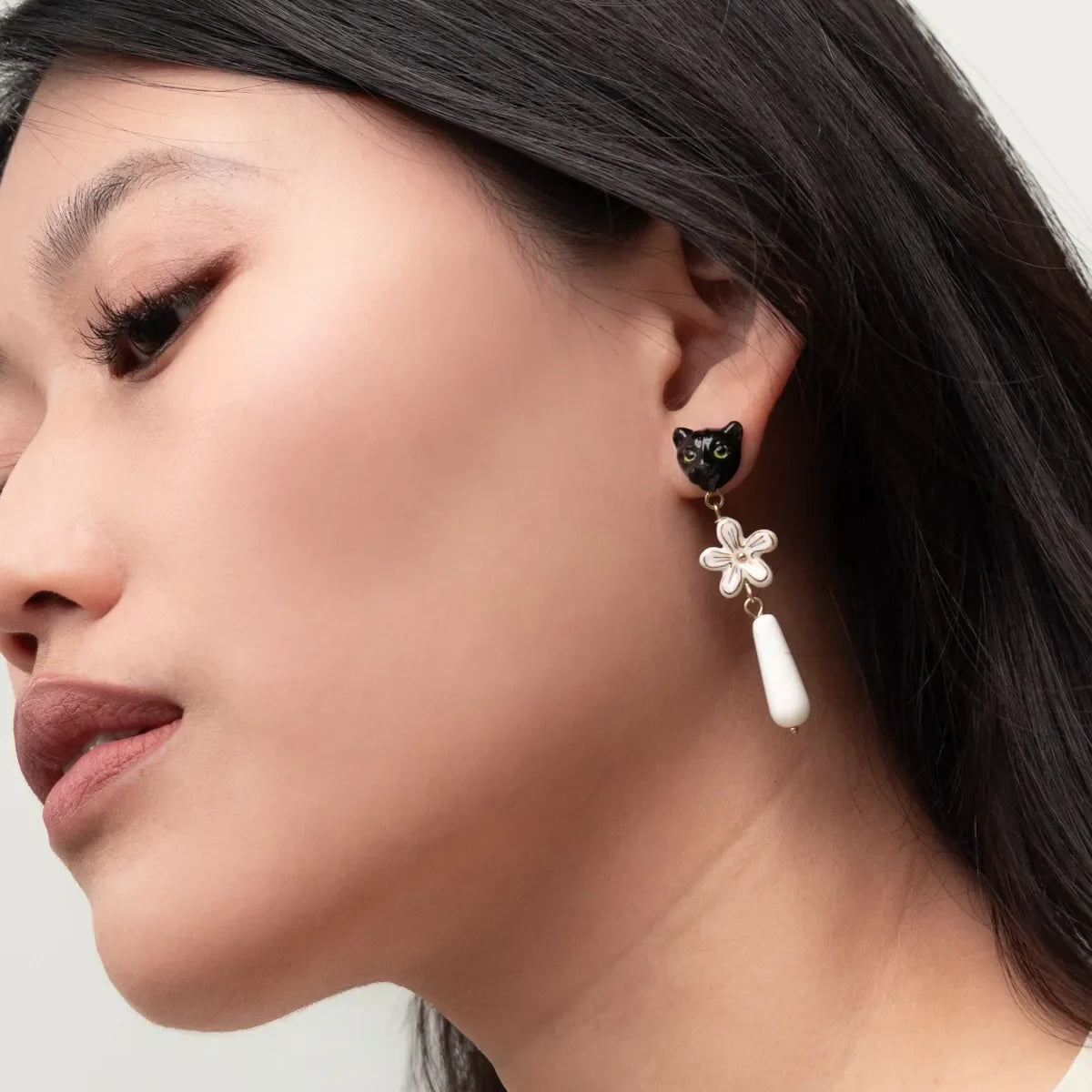 A close-up side profile of a woman wearing a dangling earring. The earring features a small black cat head charm, a white flower-shaped charm, and a white teardrop drop, shown against her dark hair and light skin.