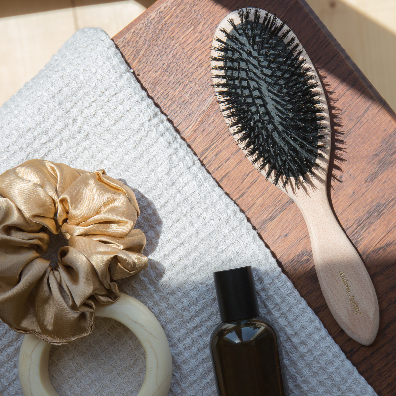 Boar bristle brush on a wooden board beside a silk scrunchie and a dark glass bottle – Andrée Jardin