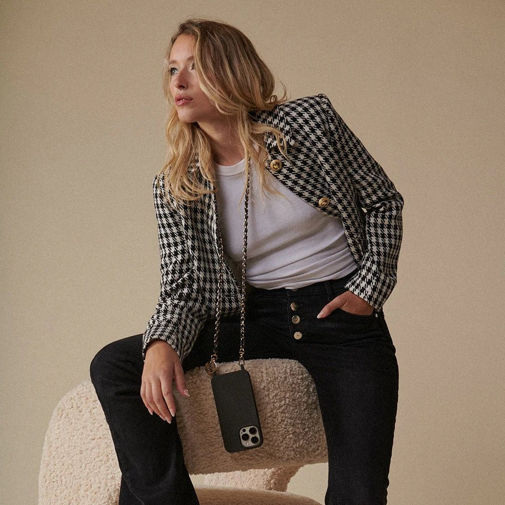 A person seated wearing a black and white checkered jacket, white top, and black trousers, accessorized with a long black and gold metal chain phone strap.