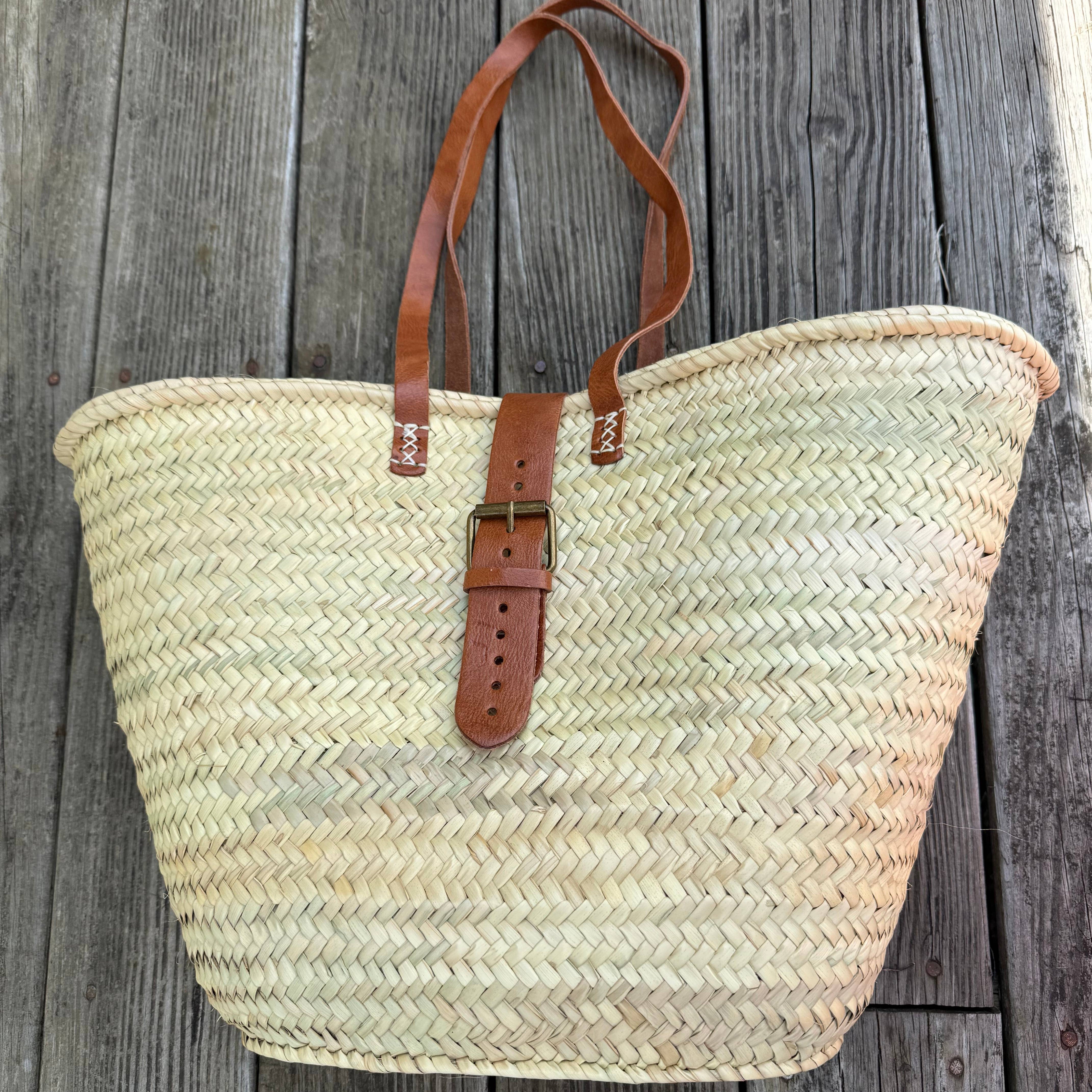 Palm leaf market basket on weathered wood, long leather handles laid across the front – Medina
