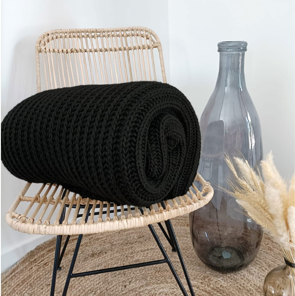 Black knitted blanket on a wicker chair with a vase and dried plants in the background