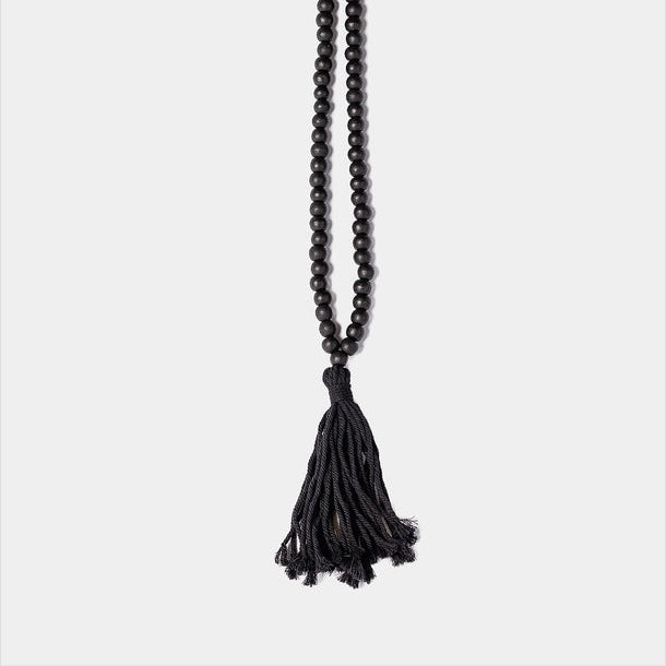 A long strand of small black round beads with a large black tassel at the end, photographed against a plain light gray background.