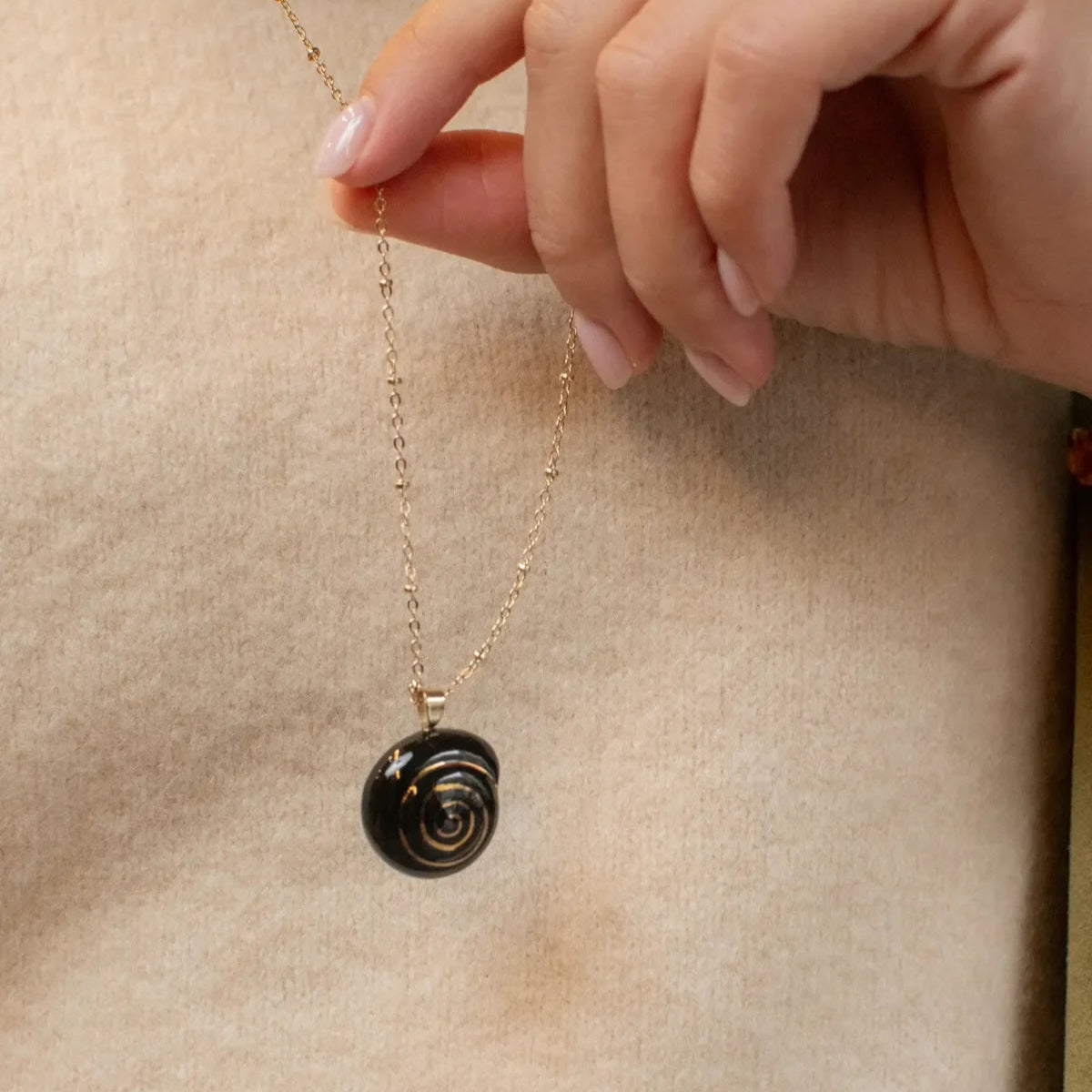 Hand holding a necklace with a black spiral pendant on a beige background