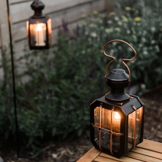Two lit lanterns on a wooden surface with a garden background