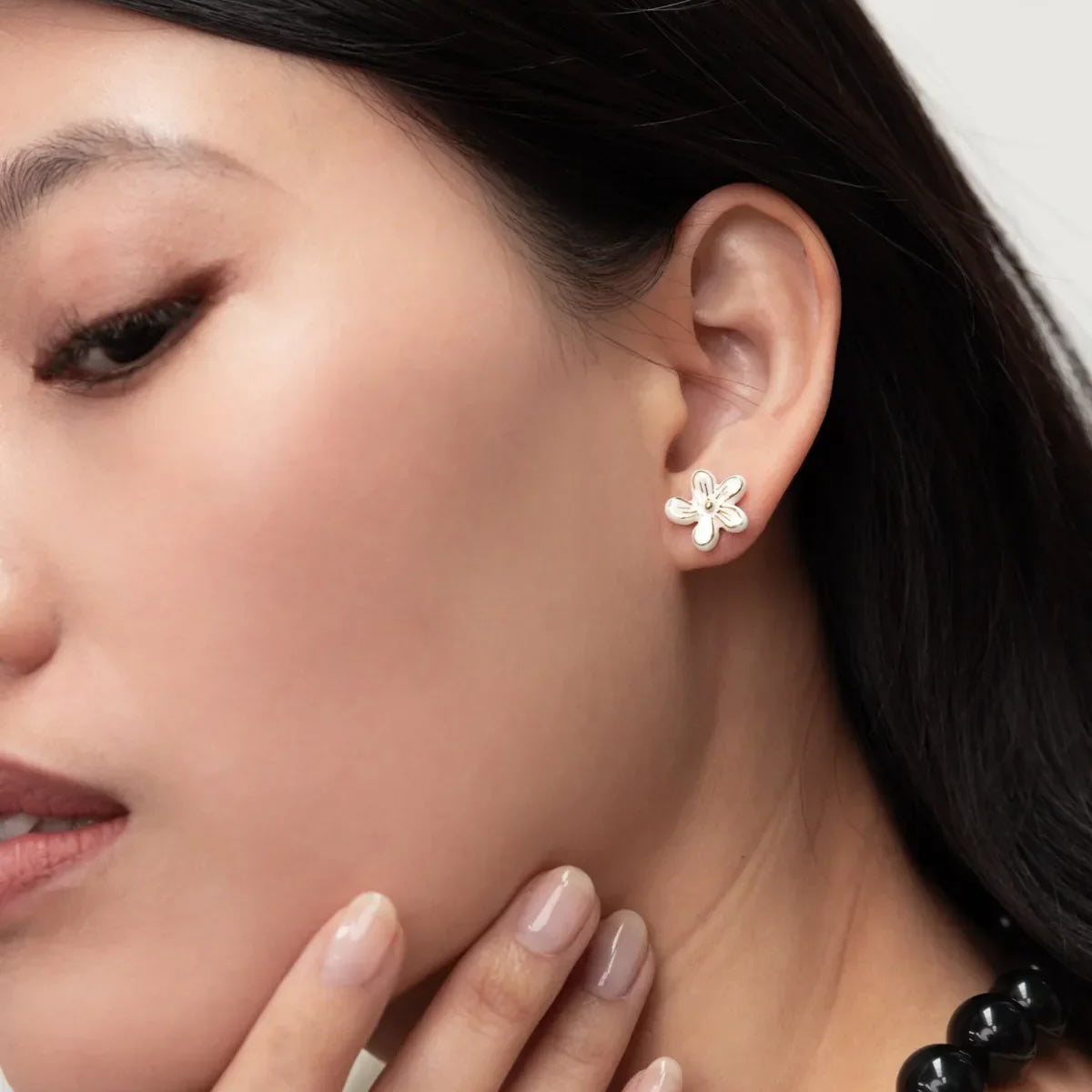 A close-up of a woman's ear wearing a small silver flower-shaped stud earring, with her hand near her chin and a black beaded necklace visible against a light neutral background.