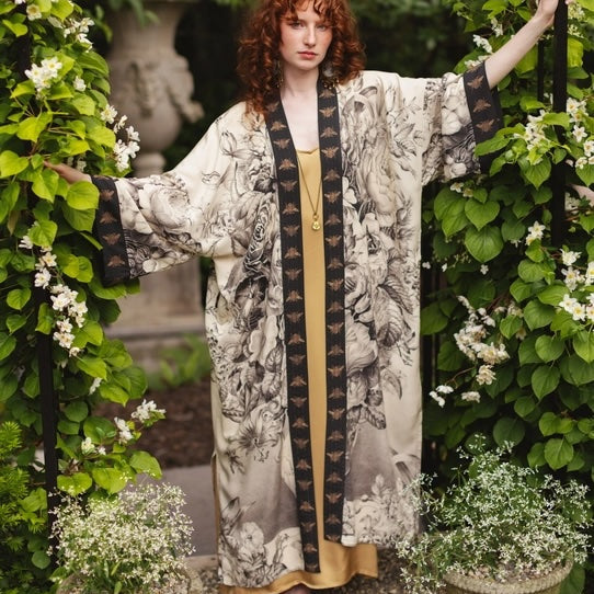Woman in a floral kimono standing in a garden with greenery and flowers.