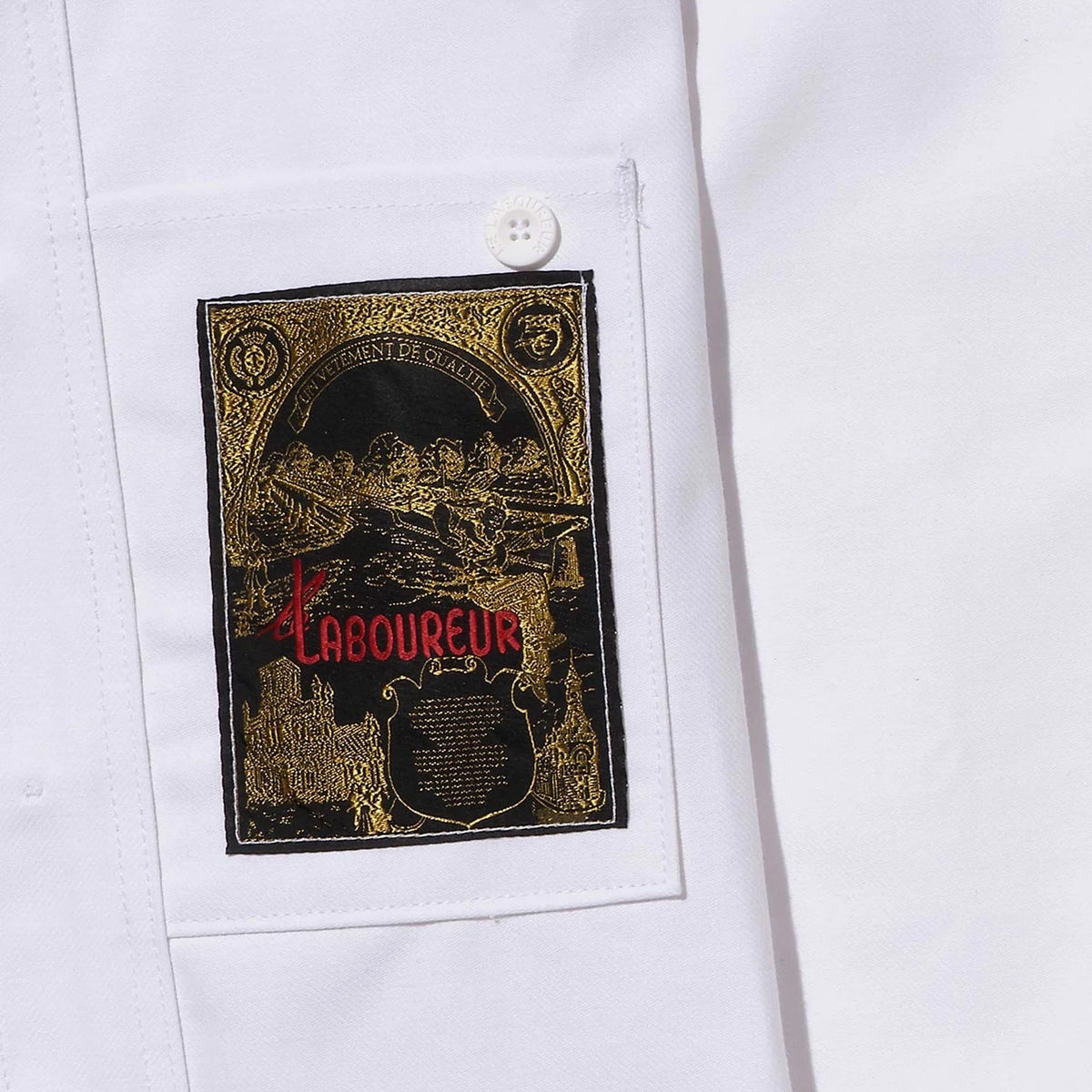 Close-up of a white fabric garment featuring a rectangular black and gold patch with red 'LABOUREUR' lettering, secured with a white button.