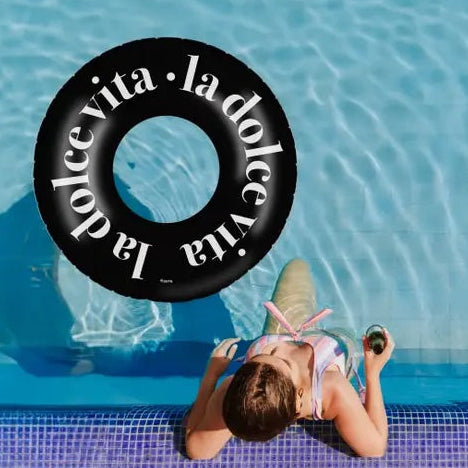 A black inflatable pool float with white 'la dolce vita' text floating above a child lying on the edge of a blue swimming pool.