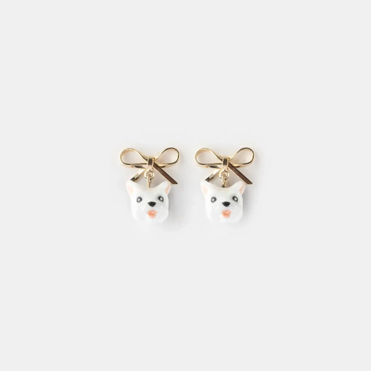 A pair of gold-tone stud earrings featuring white cat faces with black eyes and noses, each adorned with a small bow on top, shown against a light gray background.
