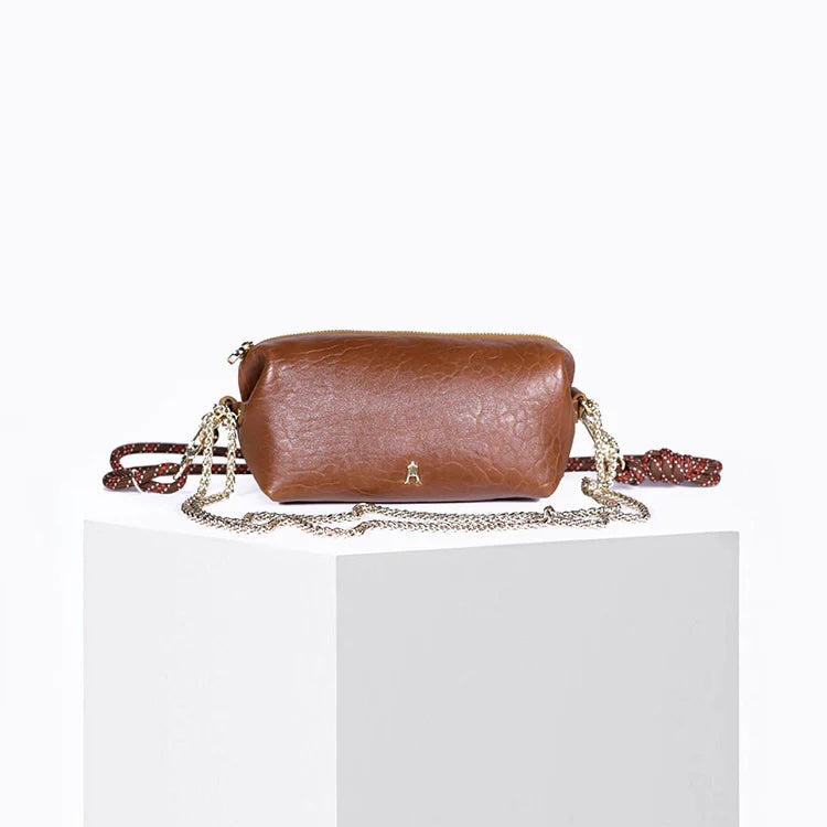 A brown textured leather shoulder bag with a silver chain strap and a red woven accent, displayed on a white block against a white background.