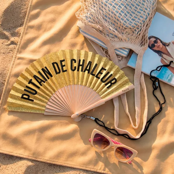 A gold glitter folding fan with black text reading 'PUTAIN DE CHALEUR' is laid on a beige blanket outdoors. A beige mesh tote bag, pink sunglasses, and a magazine are also arranged around it on sand.