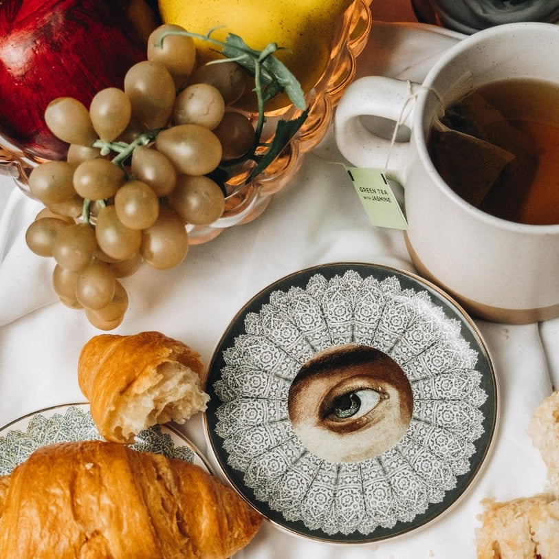 Tea cup with a saucer featuring an eye design, croissants, grapes, and apples on a white surface.