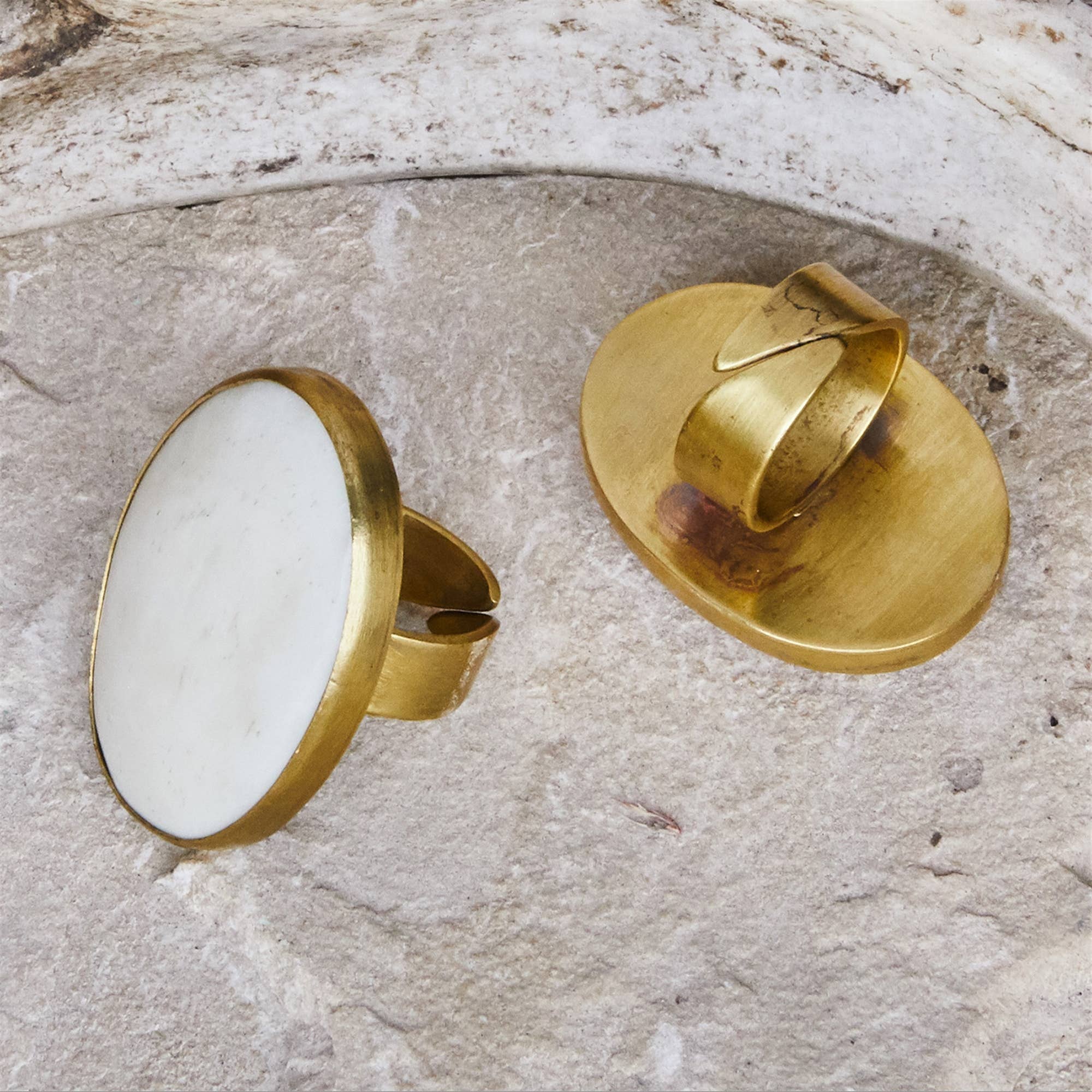 Two vintage-style gold rings displayed on a textured stone surface. One ring features a large, flat oval white stone, while the other shows the back of the band with its setting.