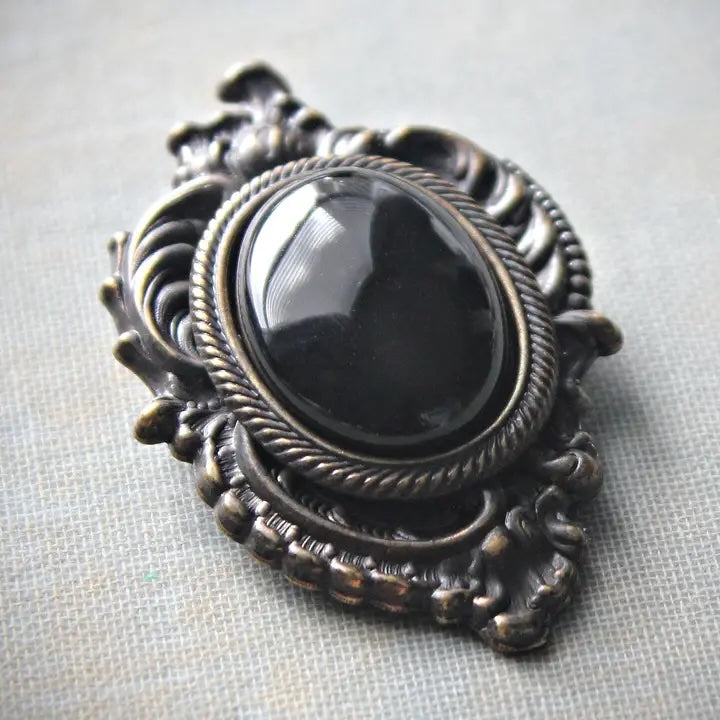 An ornate oval brooch featuring a glossy black cabochon stone surrounded by a rope-style border and an intricate antique bronze filigree frame, set against a textured light gray fabric background.