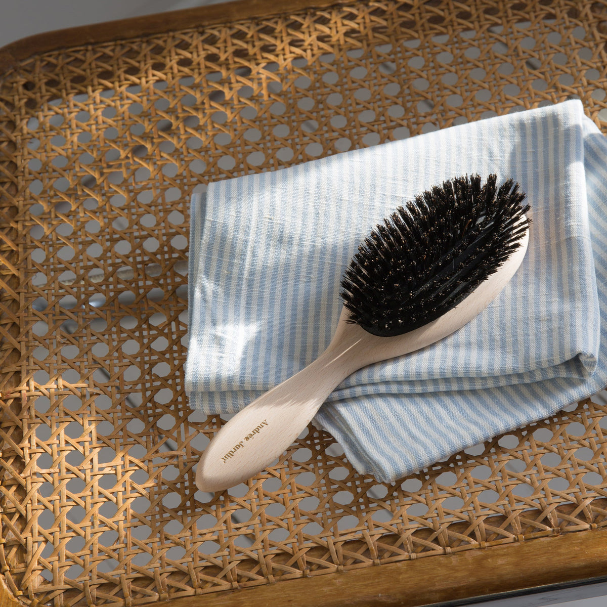 Oval beech hair brush with black boar bristles resting on striped linen on a caned tray – Andrée Jardin