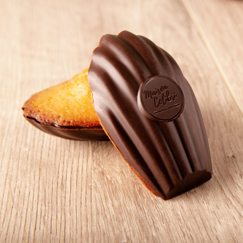 Close-up of a dark chocolate coated madeleine on a rustic wooden table, golden cake peeking from its shell – Maison Colibri