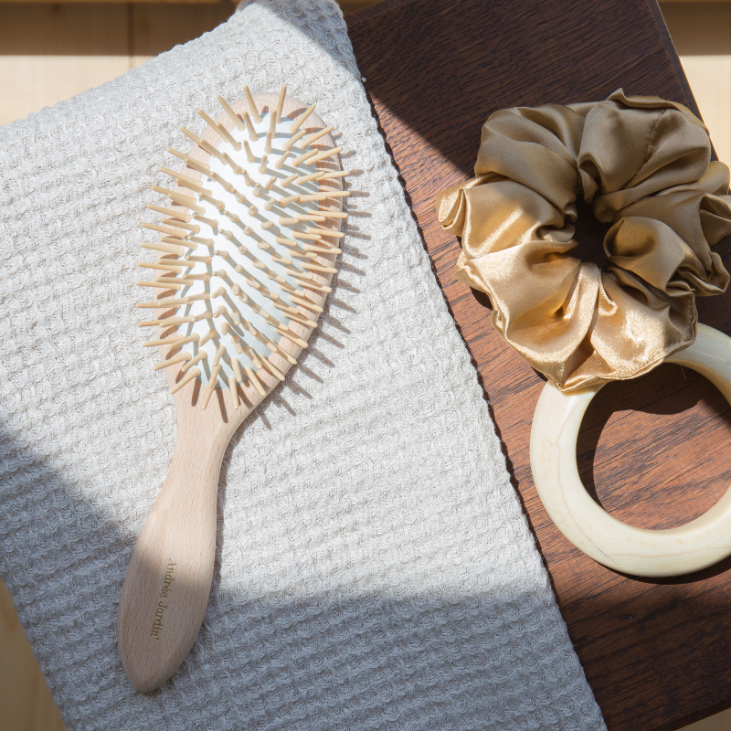 Wooden-pin detangling brush on a wooden board beside a gold silk scrunchie – Andrée Jardin