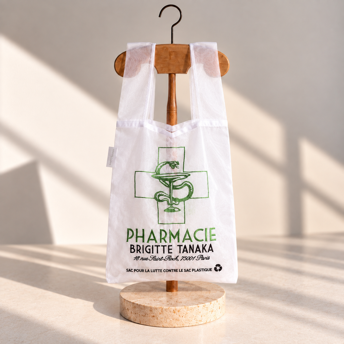 Organza shop bag embroidered with a green pharmacy cross and serpent, on a wooden hanger – Brigitte Tanaka