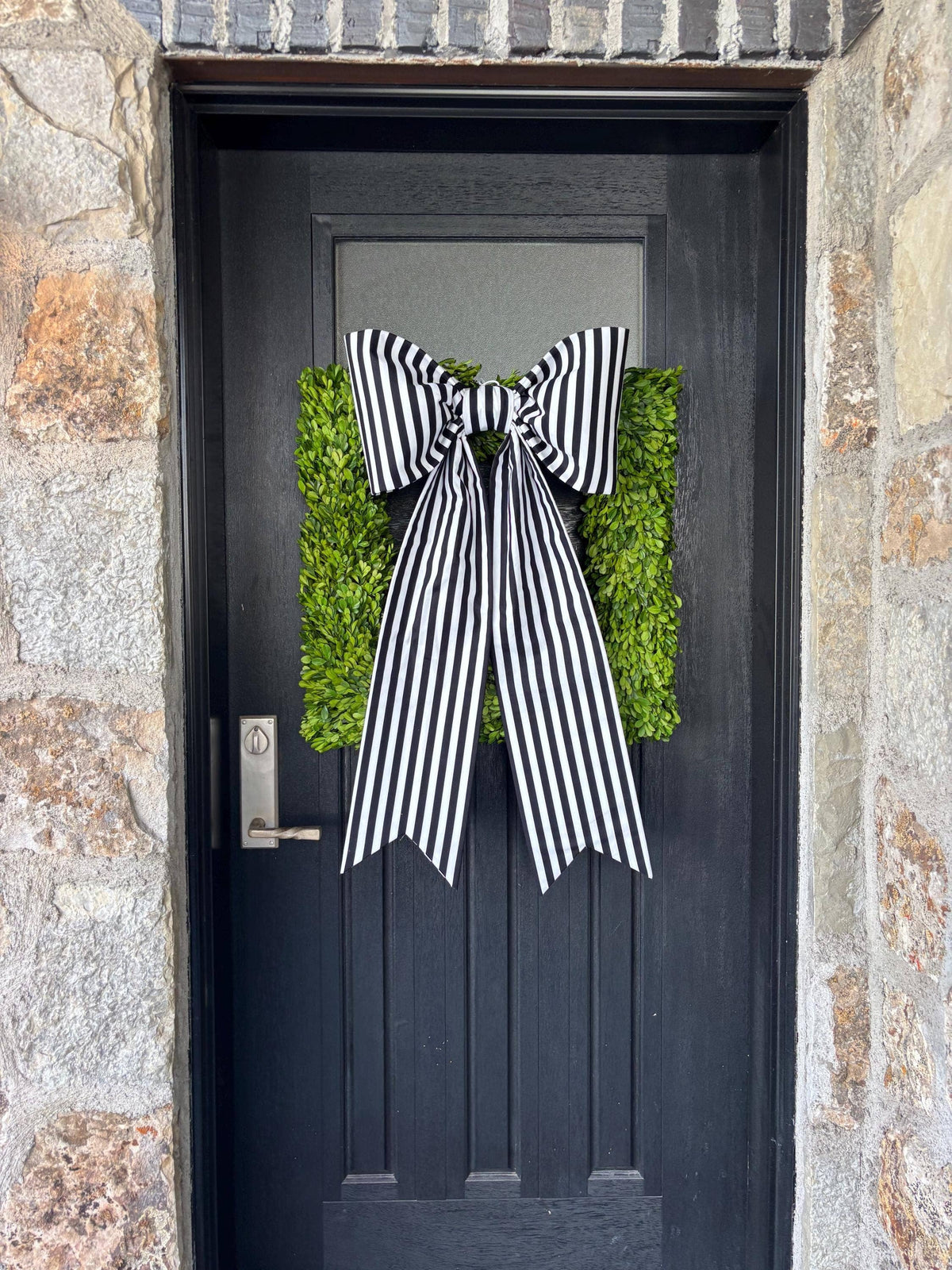 Large black and white striped bow on a boxwood wreath hung on a dark front door – La Maison Reverie