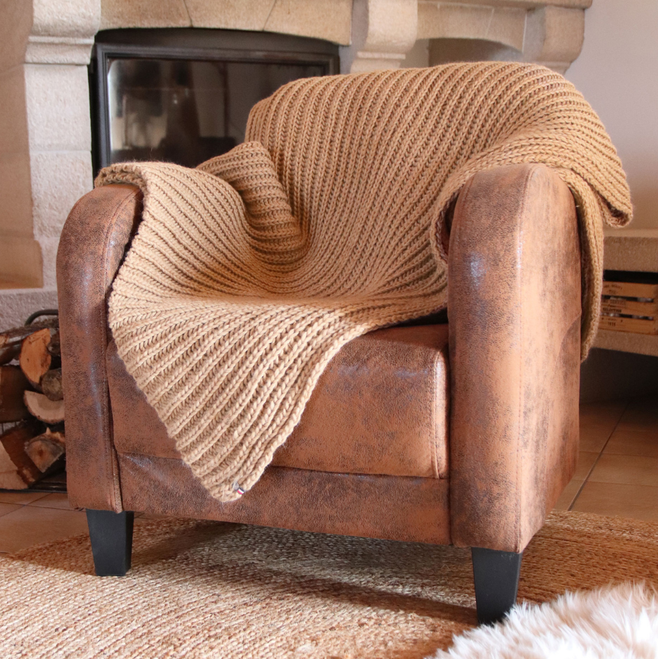 Brown leather armchair with a beige knitted blanket draped over it, in front of a fireplace.