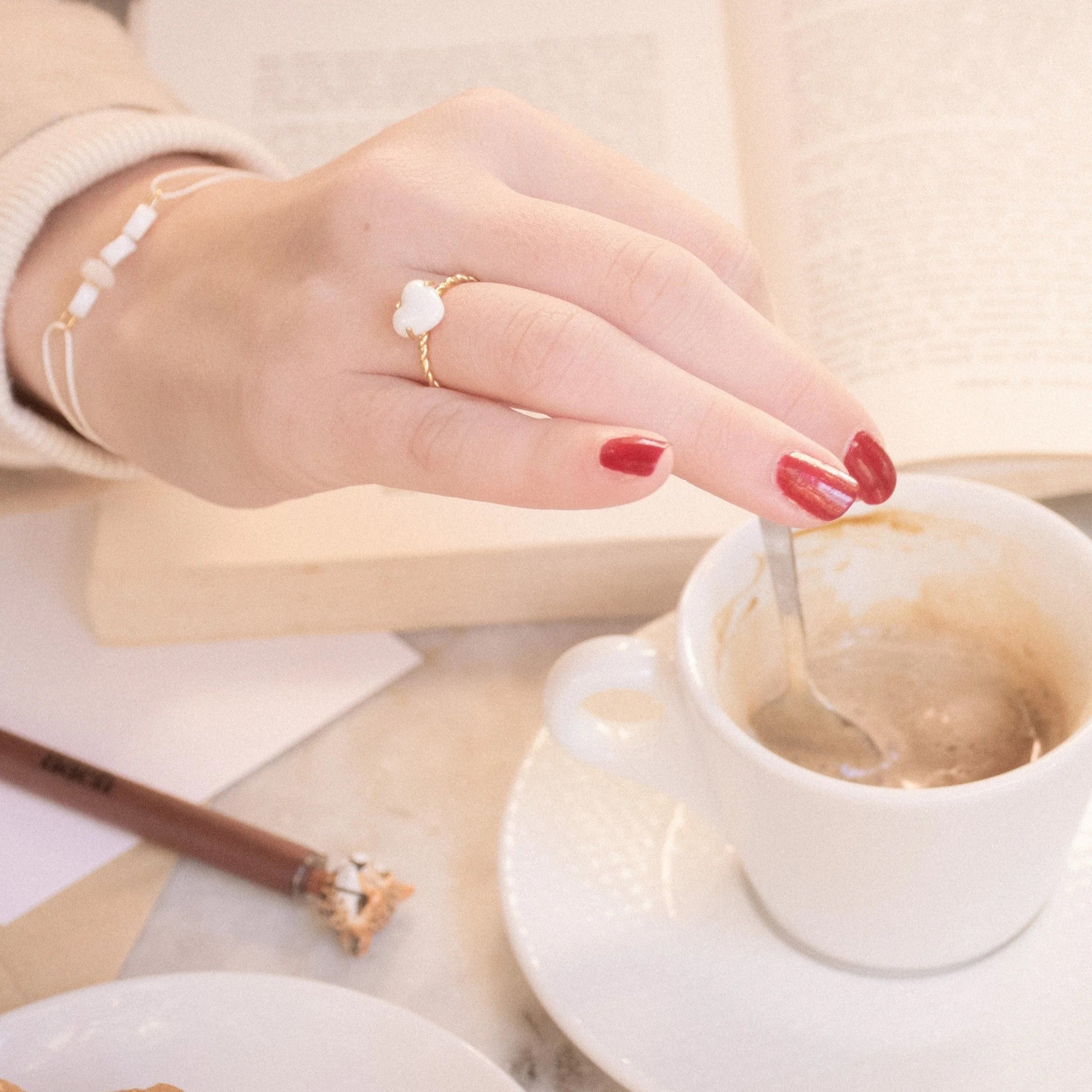 Hand stirring coffee with a spoon, wearing a ring, on a table with a book and pastry.