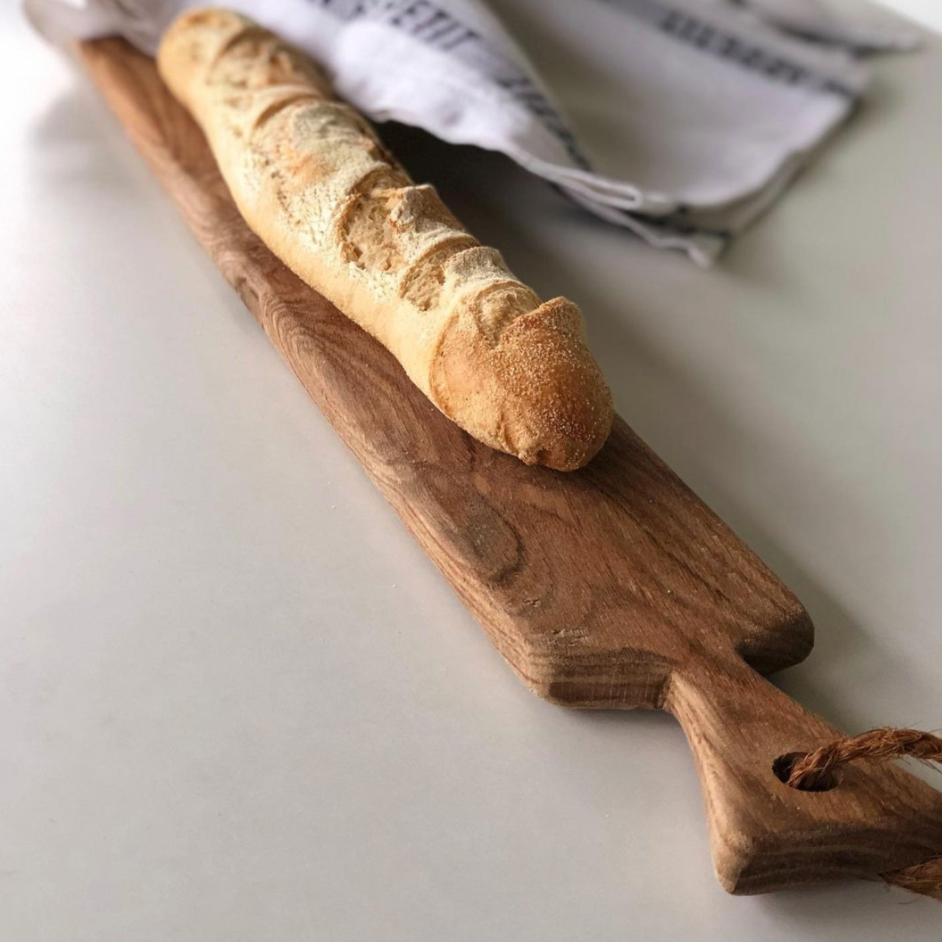 A long, rustic oak cutting board with a freshly baked baguette on top, placed on a white surface with a checkered napkin at the side.